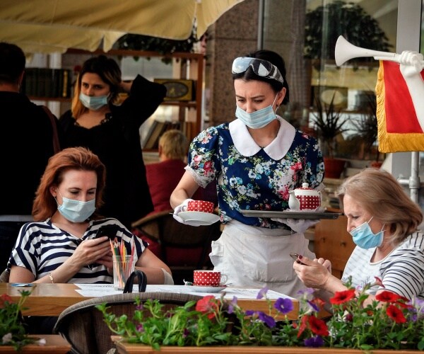 waitress in a blue floral top and face mask serves two women sitting down and wearing masks