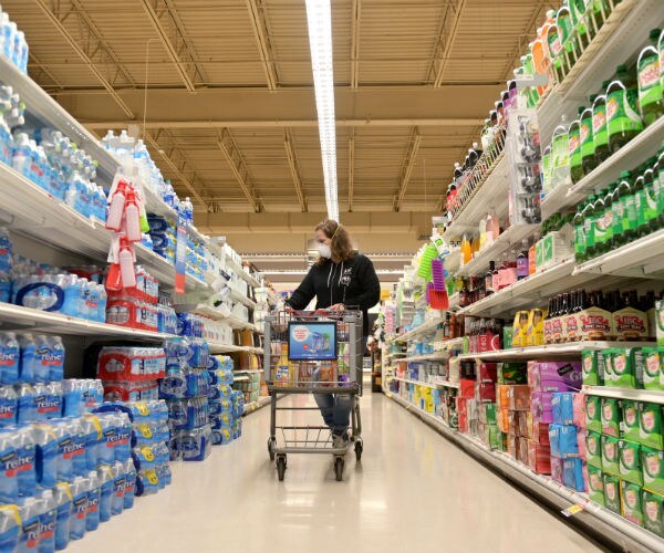 a woman, with a white facial mask on,  is shopping in the middle of a soda and bottled water aisle in a grocery store.