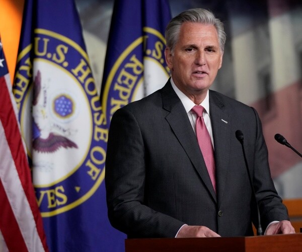 mccarthy in a dark gray suit and pink tie in front of flags with the house of representatives seal