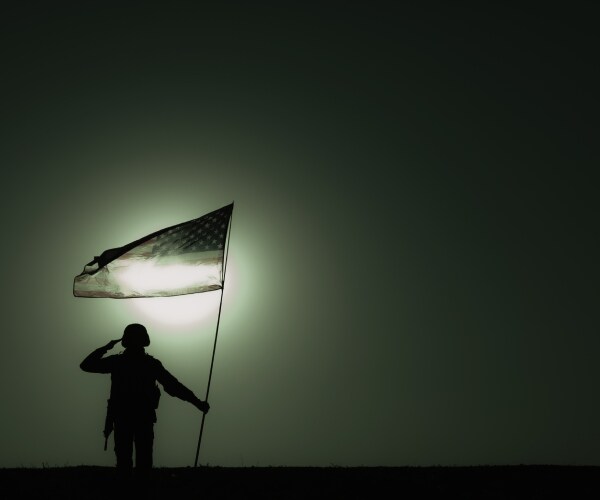 one soldier holding an american flag and saluting in a barren area