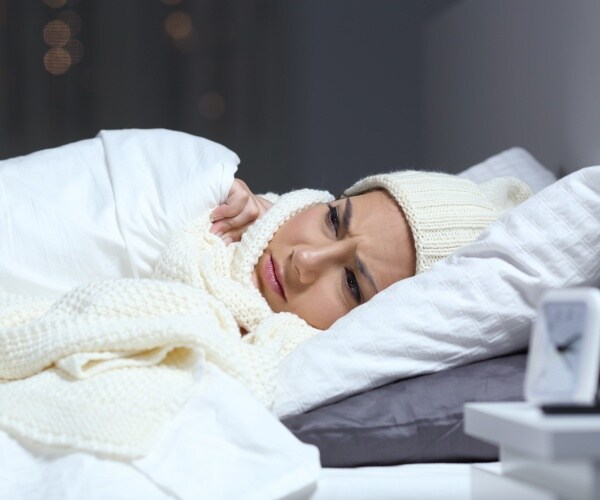 woman in bed in white comforter, white hat and scarf looking cold
