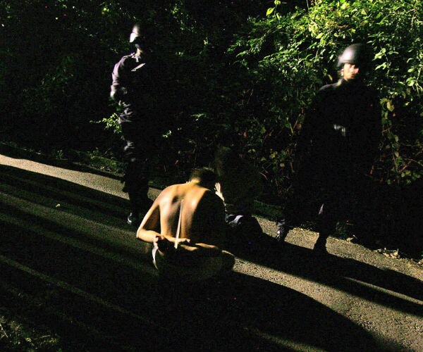 a shirtless man kneels before two armed men wearing hard hats