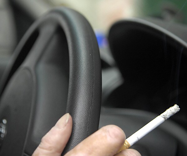 a driver grips the wheel with his right hand while holding a lit cigarette between his index and middle finger