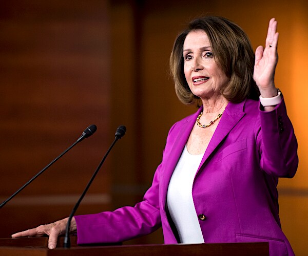 nancy pelosi raises her left hand while speaking to the media before a podium and a gaggle of mics.