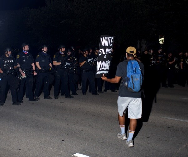 black man holding a sign in protest stands before a row of police officers