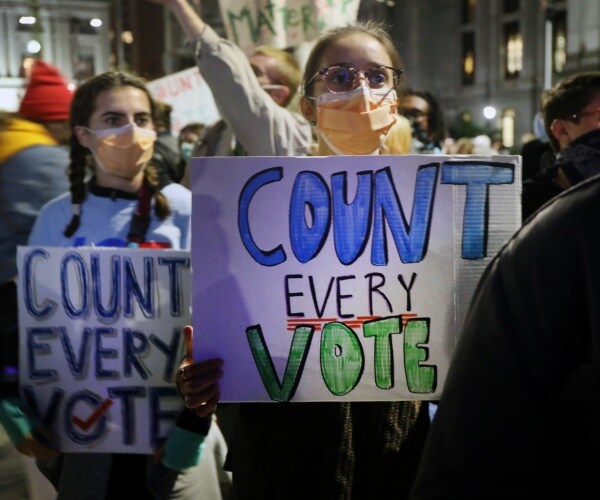 women, wearing facemasks,  hold signs saying count every vote amongst a crowd of people
