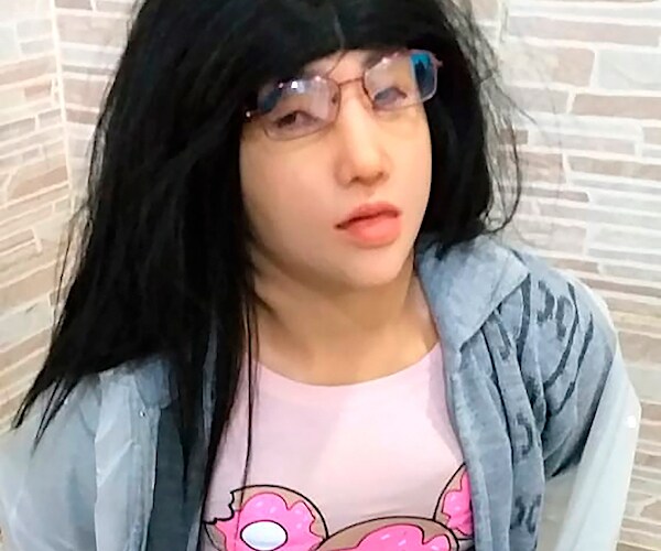 a man wearing a mask, wig, glasses, and feminine clothing, has his hands confined behind his back