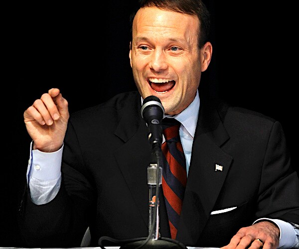 adam andrzejewski gestures with his right hand while delivering remarks into a mic