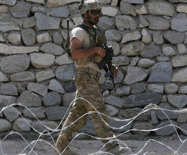 an american soldier paroles in front of a rock wall and behind barbed wire