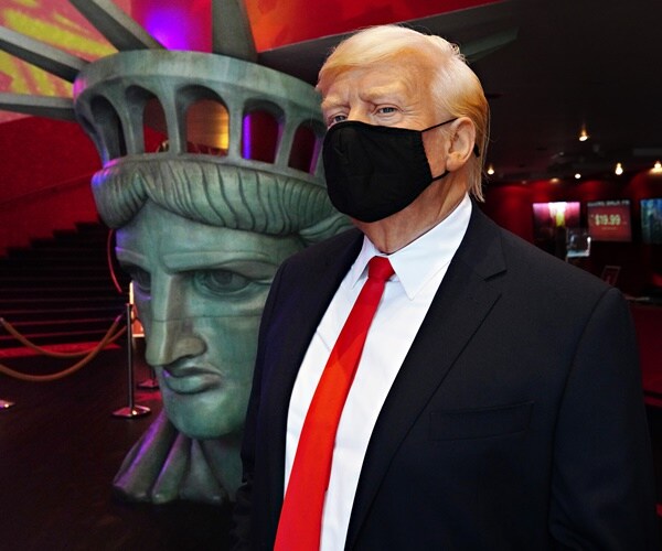 a maksed wax figure of donald trump stands in front of a lady liberty head
