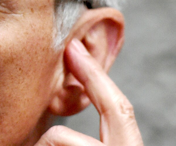 a gray haired man points to his left ear with his left index finger