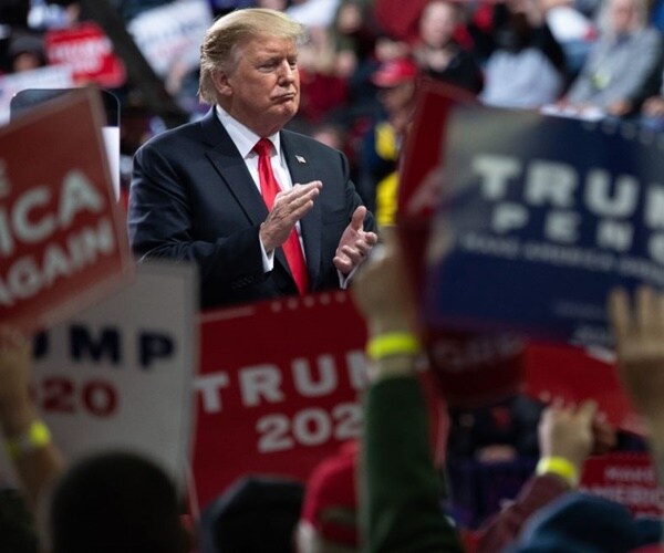 donald trump stands on a stage at a rally surrounded by signs from supporters