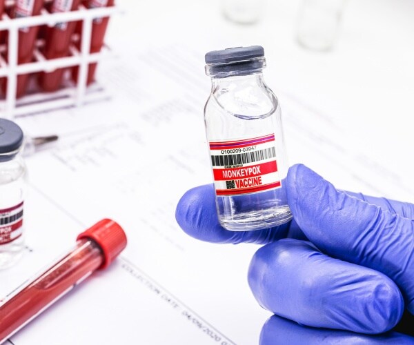 hand with surgical glove holding up a vial that says 'monkeypox vaccine', syringe and more doses on table