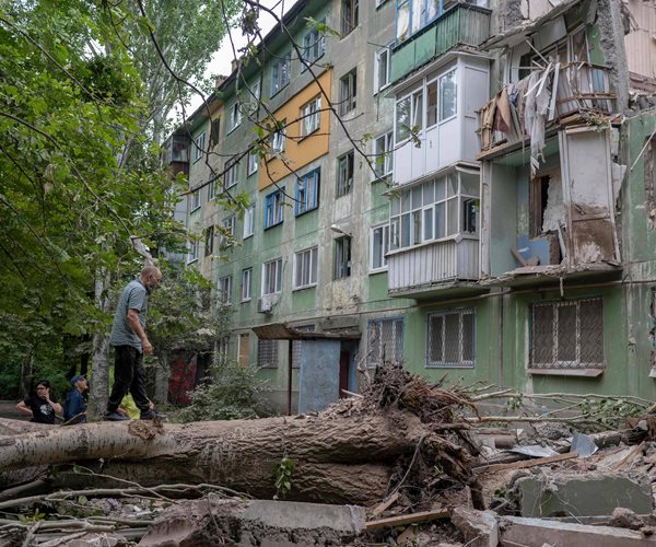 Three people on the ground and one walking on a felled tree look at debris and the bombed facade of a building.