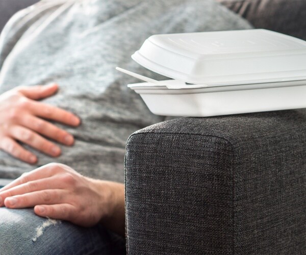 man sitting in chair with a take out food container next to him, holding stomach because not feeling well