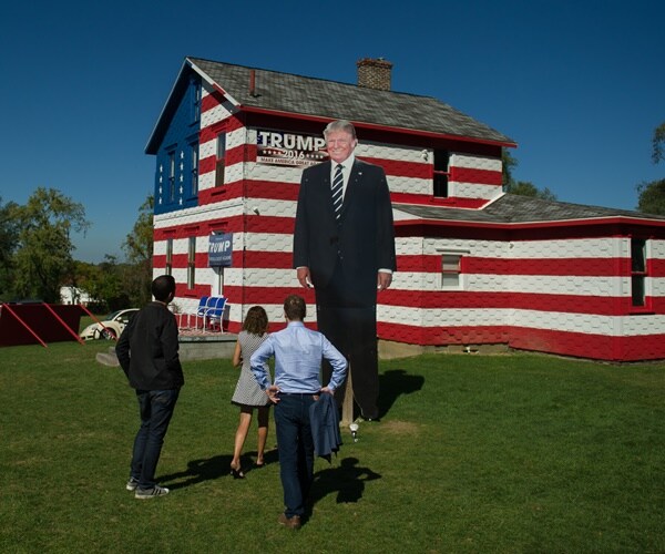 people stand in front of a red, white and blue painted house with a giant cardboard donald trump