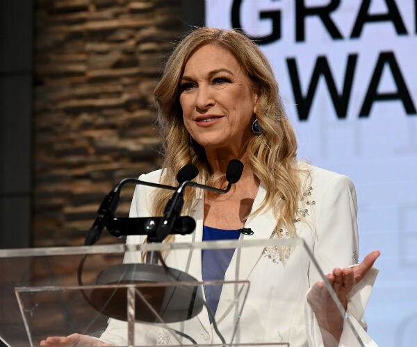 deborah dugan is shown on stage at a podium wearing a blue shell under a white suit coat