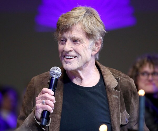 redford in a brown jacket and black t shirt holds a mic