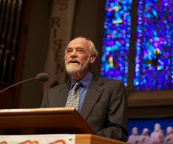 eugene peterson speaks in front of stained glass church windows and behind a podium mic