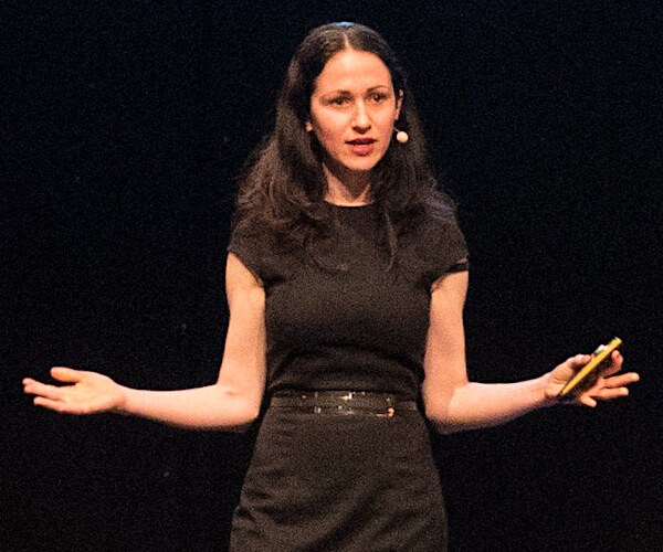 a women stands and delivers a speech on stage at a conference, extending both arms out to her side with palms up