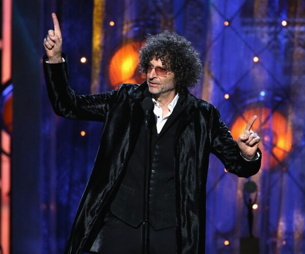howard stern in a black suit and black jacket standing on stage pointing his fingers up