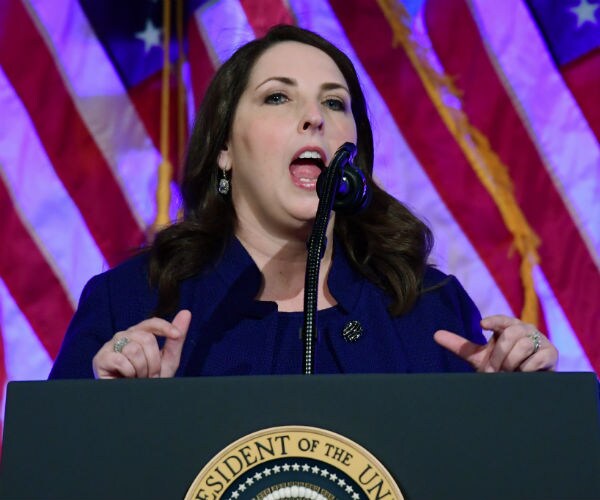 ronna mcdaniel is shown speaking behind a podium in a blue suit with the american flag draped in the background