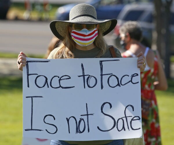 a teacher wearing a teal, pink, white and yellow mask hold a signs that reads face to face is not safe