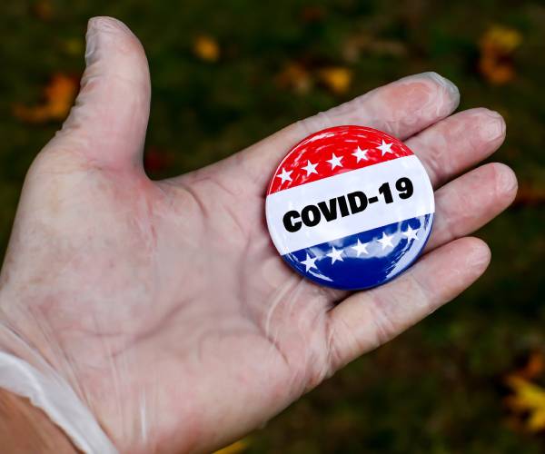 gloved hand holding a political button with covid nineteen written on it