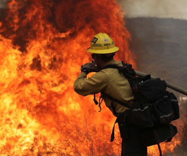 firefighter stands with a hose wearing a hardhat and backpack in front of the wildfire