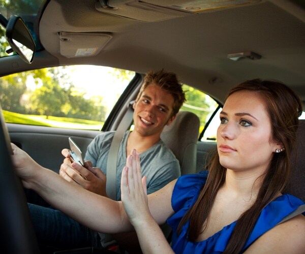 teenage girl driving puts hand up to distracting teenage boy holding up his phone to her