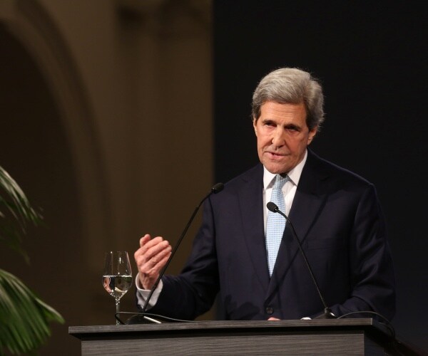 john kerry in a suit and light blue tie speaking at a podium with a glass next to him