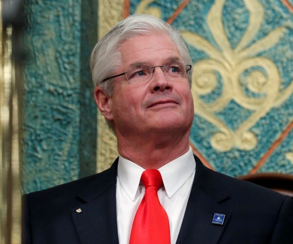 Shirkey in a suit and red tie with blue and green patterned wall behind him