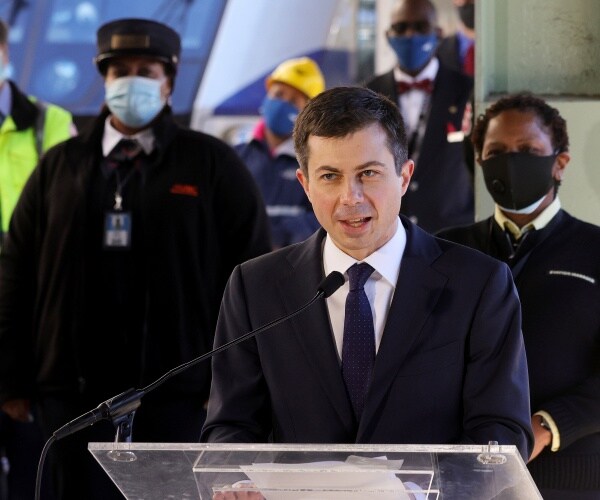 pete buttigieg speaks at a podium in front of masked people and an amtrak train