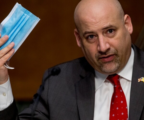 carpenito wearing a dark gray suit and red patterned tie holding up a face mask