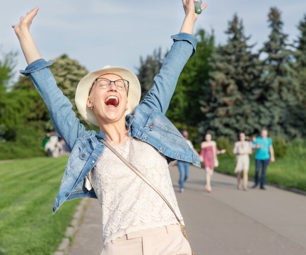 woman with no hair and a hat after chemo treatment walking on road and smiling/happy