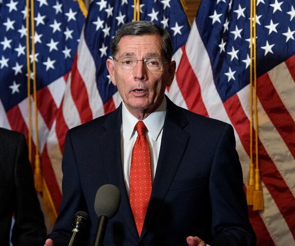 john barrasso stands in front of flags and speaks at a mic.