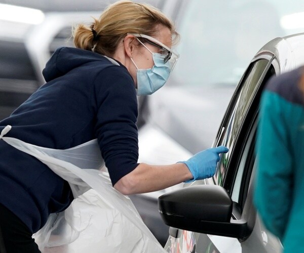 nurse wearing a navy blue hoodie and plastic protective suit takes a swab of a person inside a car