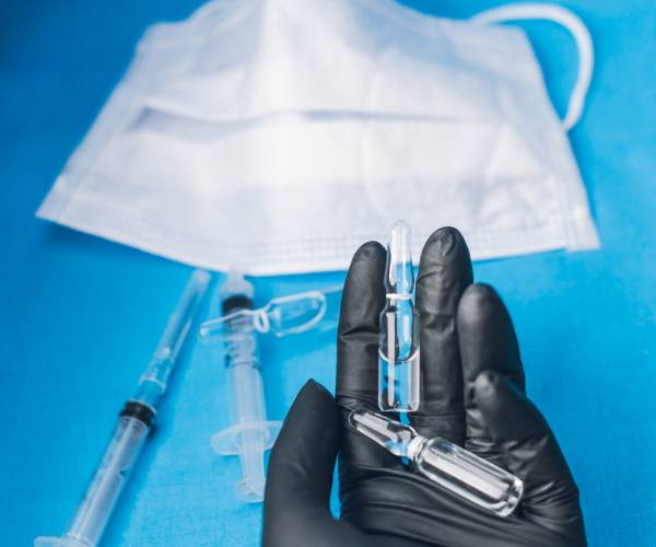 gloved hand holding vials with a mask and syringes in the background