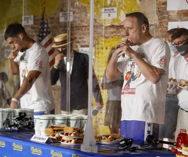 two men eating hotdogs with staff members standing nearby wearing face masks