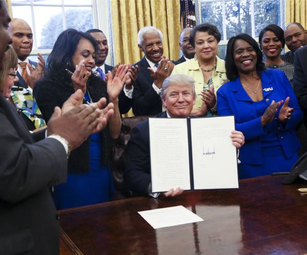 president trump sitting at his desk holding a signed document surrounded by people standing behind him