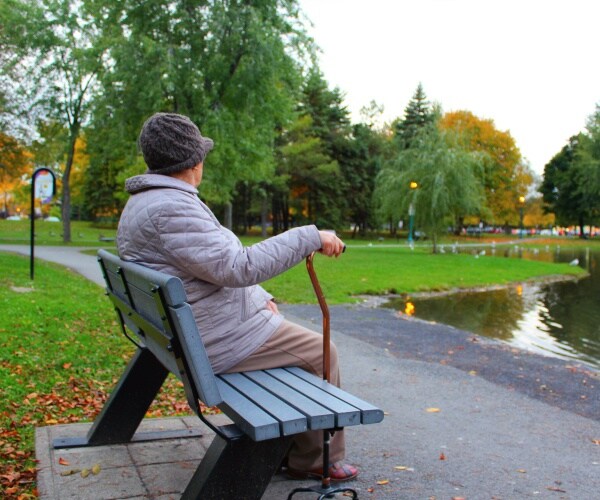 older woman sitting on bench in park by herself looking out at ducks on pond
