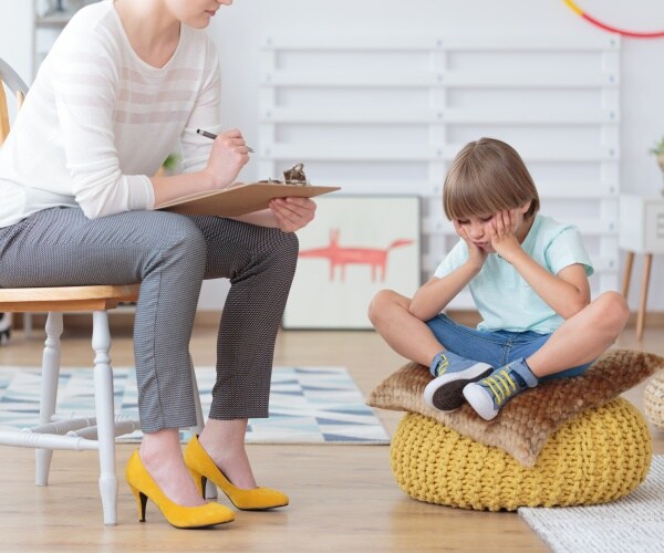young boy sitting on low pillow looking sad and anxious, a psychologist with a clipboard talking to him