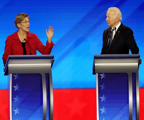 warren in a red blazer and black top speaking as biden in a black suit listens on the debate stage