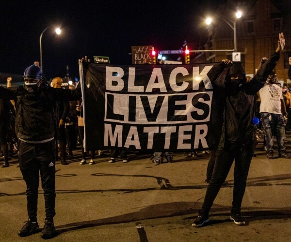 two men dressed in black hold up a blm flag with other protesters behind them
