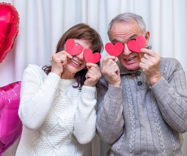 older couple smiling and holding pink/red hearts over eyes, with two heart-shaped ballons