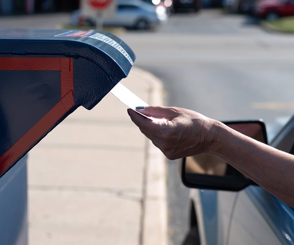 A woman sitting in the driver's seat reaches through a car's open window and inserts a mail-in ballot into a mailbox.