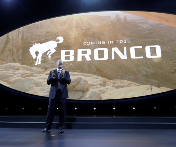 hinrichs in a dark colored suit stands on stage with a bronco logo and announcement projected behind him