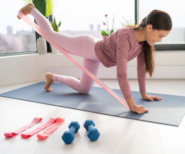 woman in pink leggings and top uses an exercise band on a mat with dumbbells and bands beside her