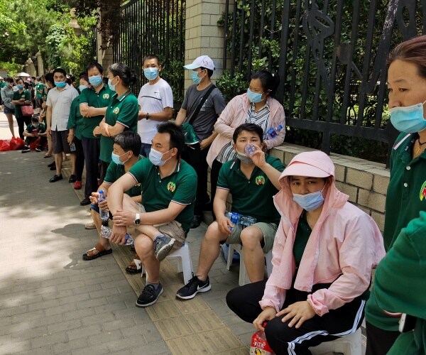people wearing face masks and green or pink shirts stand and sit in line outside