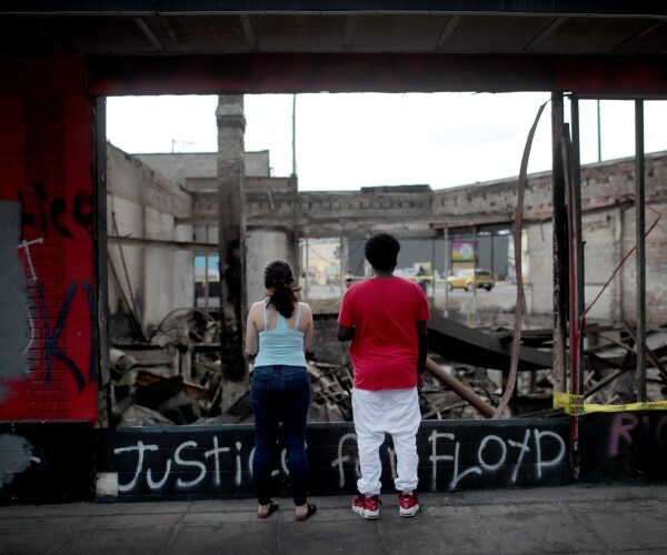 two young people stand in front of wreckage that was burned in a riot with justice for floyd spray painted below